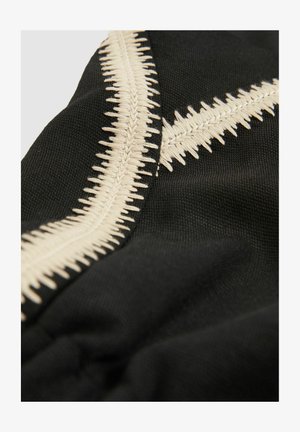 Tissu noir ornée de broderies crème complexes avec des motifs en zigzag, mettant en valeur une surface texturée et des points détaillés.