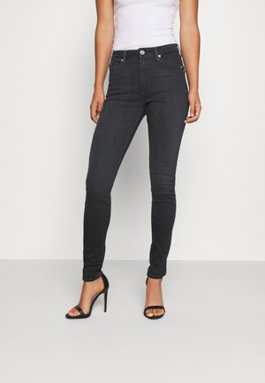 Vrouw die een zwarte skinny jeans met hoge taille, een wit mouwloos topje en zwarte open-teen hoge hakken draagt, staand tegen een effen achtergrond.