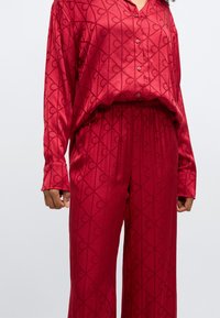 Czerwony satynowy komplet piżamowy składający się z koszuli na guziki z długim rękawem oraz spodni z elastycznym paskiem, ozdobiony geometrycznym wzorem.
