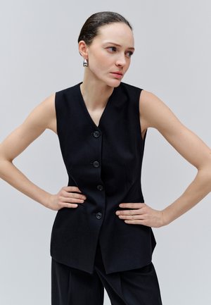 Femeie cu păr închis la culoare prins într-un coc neted, purtând un vestă neagră fără mâneci cu nasturi și pantaloni asortați, pozând cu mâinile în șolduri pe un fundal gri.