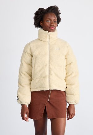 Cream-colored fleece jacket with a high collar, front zipper, and elastic cuffs. Worn over a brown mini skirt with a zip front.