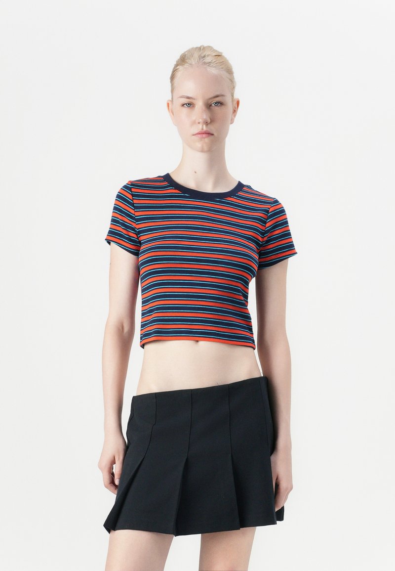 Crop top dengan garis horizontal merah, biru, dan navy, dengan leher bulat dan lengan pendek. Dipadukan dengan rok plisket hitam.