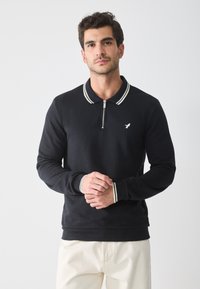 Sort langærmet polo-shirt med lynlås krave, med hvide stribede detaljer på kraven og manchetterne. Lille logo på venstre bryst.