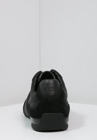 Mustank BOSS-kengät nähtynä takaa, joissa on nahka- ja mokka-aineita sekä kohokuvioitu brändilogo kantapään kohdalla.