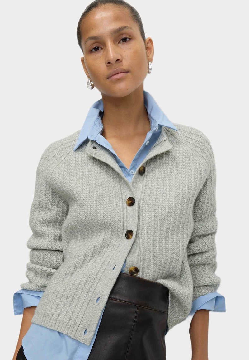 Grauer gerippter Cardigan mit braunen Knöpfen, getragen über einem hellblauen Hemd mit Kragen. Hat eine lockere Passform und ein strukturiertes Strickmuster.