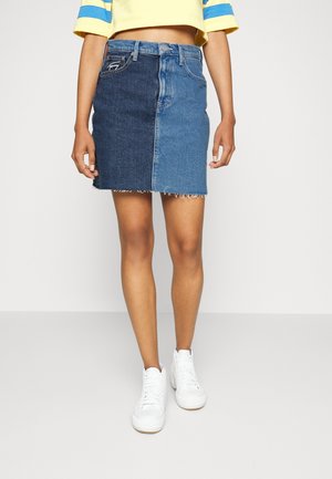 Person iført et højtaljet, tofarvet denimskørt med rå kant, en cropped gul top med blå striber og hvide sneakers.