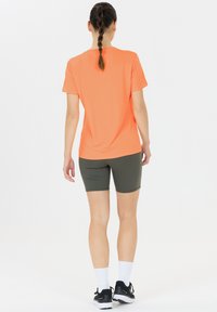 Oranssi lyhythihainen urheilu-t-paita yhdistettynä oliivinvihreisiin pyöräilyshortseihin. Mallilla on mustat urheilukengät ja valkoiset putkihuput.