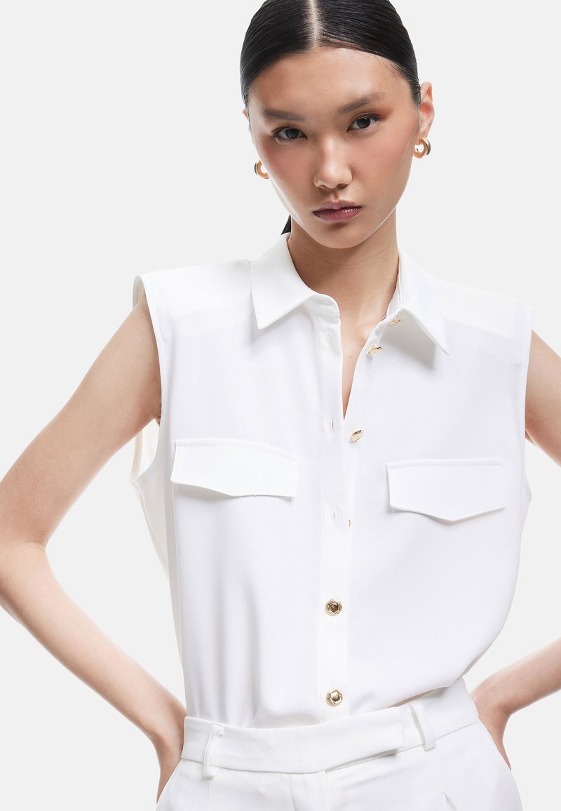 Blouse blanche sans manches avec un col, comportant deux poches poitrine, des boutons dorés et une texture lisse. Portée avec un pantalon assorti.