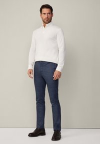 Weißer Baumwollpullover mit Kragen, kombiniert mit strukturierten marineblauen Hosen. Das Model steht mit den Händen an den Seiten und trägt braune Lederschuhe.