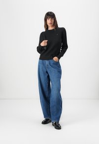 Pull en maille noire avec un motif texturé, associé à un jean en denim bleu décontracté et des chaussures noires. Le mannequin pose devant un fond blanc uni.