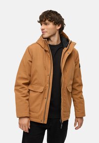 Brun yderjakke med lynlås, to forlommer og en justerbar hætte. Båret over en sort sweater af struktureret stof.