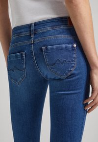 Mörkblå denimjeans med en kroppsnära passform, två bakfickor med ett distinkt vågmönster och kontrasterande sömnadsdetaljer.