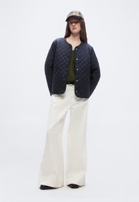 Γυναίκα φορώντας μπλε μπουφάν με καπιτονέ ύφασμα, ελιάς πράσινη μπλούζα, φαρδιά λευκά παντελόνια, μαύρα παπούτσια και καρό καπέλο, στέκεται μπροστά σε λευκό φόντο.