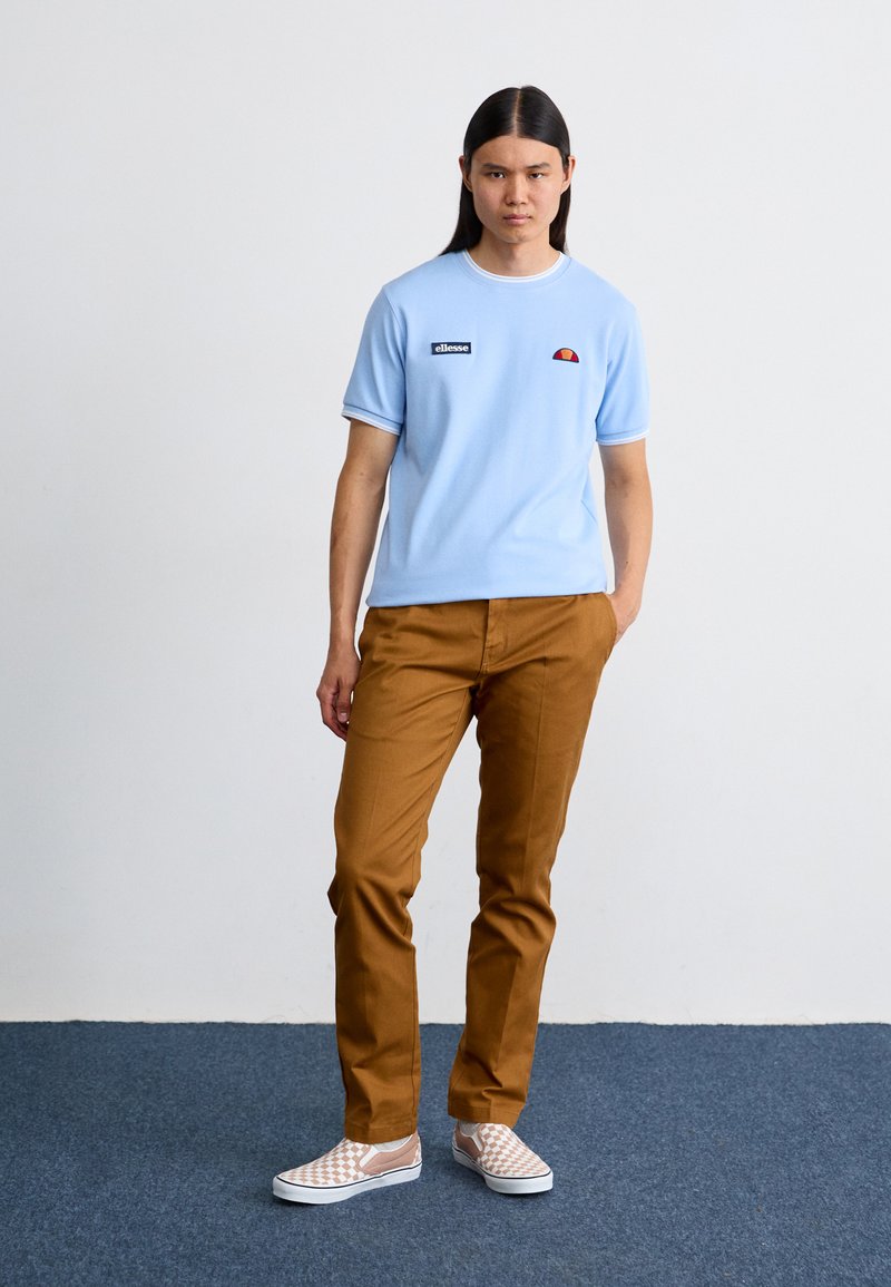 Himmelblaues kurzärmeliges T-Shirt mit einem Logo, beige gerade geschnittene Hose und weiße Slipper mit einem Schachbrettmuster.