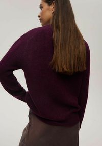 Burgundowy sweter z dzianiny o luźnym kroju, z długimi rękawami i prążkowanymi mankietami, w połączeniu z brązową spódnicą z warstw. Miękka tekstura, minimalne detale.