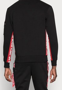 Sweatshirt noir avec des panneaux latéraux rouges présentant du texte blanc. Fait de coton, il a une coupe décontractée et des poignets côtelés pour le confort.