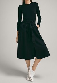 Femme portant un haut noir à manches longues et une jupe midi ceinturée assortie de baskets blanches, debout avec une jambe croisée sur l'autre.