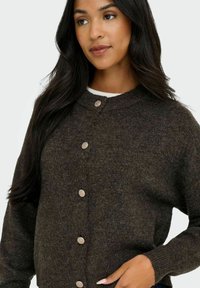 Dunkelbraune Strickjacke mit rundem Halsausschnitt, ausgestattet mit fünf großen silbernen Knöpfen und gerippten Bündchen. Weicher Stoff und oversized Schnitt.