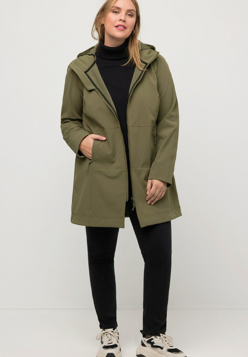 Veste à zip olive avec capuche, dotée de deux poches avant et d'une coupe décontractée. Un col roulé noir et un pantalon sombre viennent compléter le look.