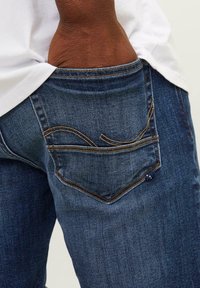 Vaqueros de mezclilla azul oscuro con un bolsillo trasero visible que presenta costuras claras y un pequeño acento metálico en el borde del bolsillo.