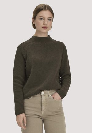 Jeune femme à la peau claire et aux cheveux courts châtain clair, portant un pull vert olive foncé et un pantalon beige, debout avec une main dans la poche.