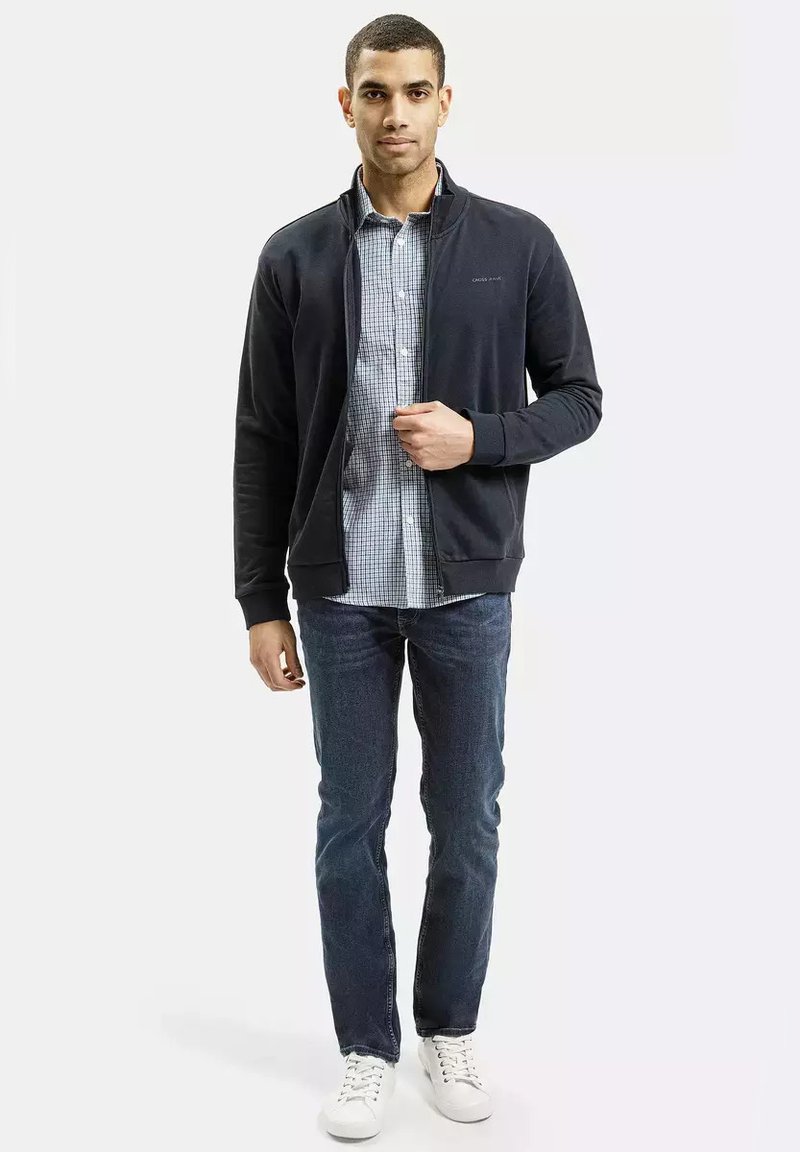 Czarna kurtka z zamkiem na plecy na jasnoniebieską koszulę w kratę, w zestawieniu z ciemnymi dżinsami i białymi trampkami. Gładki materiał i dopasowany krój.