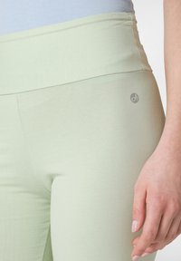 Ljusgröna träningstights med hög midja, slät yta och en liten grå logotyp på sidan. Närbild av midjan och handen.