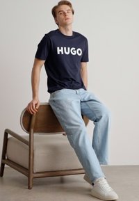 Tricou din bumbac bleumarin cu text alb "HUGO", asortat cu blugi din denim bleu deschis și adidași albi, așezat pe un scaun din lemn, pe un fundal neutru.