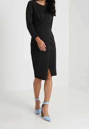 Femme portant une robe noire à manches longues longueur genou, associée à des talons hauts bleu clair à bride à la cheville, debout dans un espace blanc simple.