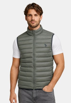 Man with light brown hair wearing gray quilted zip-up sleeveless vest over white short-sleeve t-shirt and gray jeans, hand in pocket.