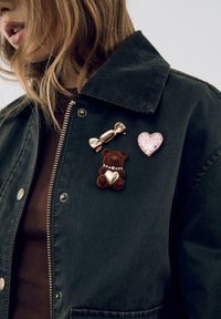 Czarna kurtka z kordurowej tkaniny z trzema przypinkami: złotą przypinką w kształcie cukierka, pluszową brązową przypinką w kształcie misia z serduszkiem oraz różową przypinką w kształcie serca obszytą cekinami.