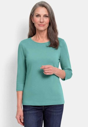 Femme aux cheveux gris portant un t-shirt à manches longues bleu sarcelle et un jean bleu foncé, debout devant un fond clair uni.