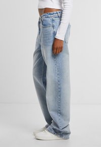 Světlé modré, vysoce pasové volné džíny s uvolněným střihem, které mají klasický pětikapsový design a mírně roztrhané lemy. Kombinováno s bílými teniskami.