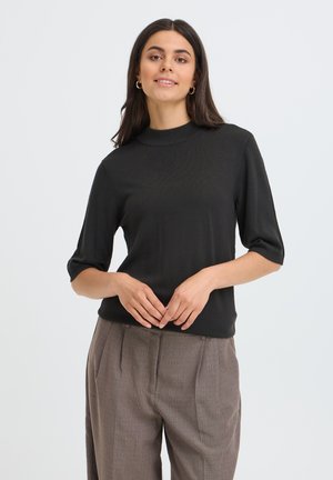Femme aux longs cheveux foncés portant un pull noir à manches courtes et un pantalon marron ample, debout les mains jointes devant elle.