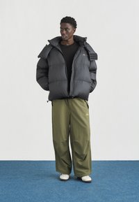 Άνδρας που στέκεται σε μπλε χαλί, φοράει μαύρο μπουφάν puffer, μαύρο πουκάμισο, χακί παντελόνι με φαρδιά μπατζάκια και λευκά αθλητικά παπούτσια, μπροστά σε λευκό τοίχο.