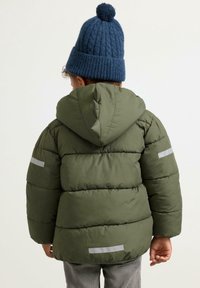 Bambino che indossa un giubbotto imbottito verde oliva con strisce riflettenti e un cappello di lana blu con pompon, di spalle contro uno sfondo semplice.