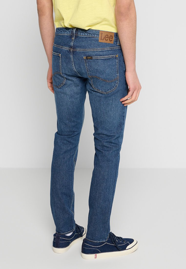 Niebieskie dżinsy o dopasowanym kroju, z średnim stanem. Z tyłu znajdują się dwie kieszenie oraz brązowa skórzana naszywka z logo na pasie.