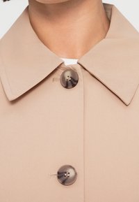 Col de manteau beige avec deux grands boutons ronds marbrés fixés sur un vêtement blanc en dessous.