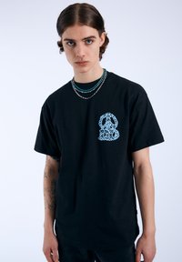 Camiseta de algodón negra con un gráfico azul de un perro en el pecho izquierdo, mangas cortas y un ajuste relajado. Presenta collares en capas.