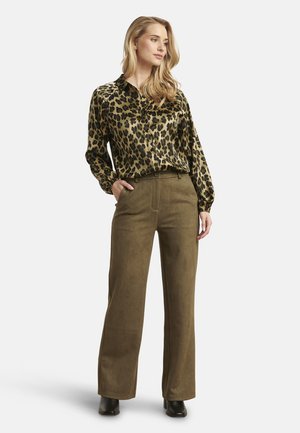 Donna con capelli biondi che indossa una camicetta a maniche lunghe con stampa leopardata, pantaloni verde oliva a vita alta e gamba larga, e stivali neri, in piedi con una mano in tasca.