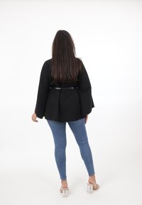 Svart, strukturerad jacka med klockärmar, åtsittande midja med ett glansigt bälte, kombinerad med blå skinny jeans och klara blockklack-sandaler.