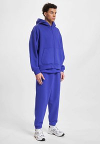 Plava dukserica sa zatvaračem sprijeda i prednjim džepovima, uparena s matching trenirkama, napravljena od mekog materijala. Model nosi bijele atletske tenisice.