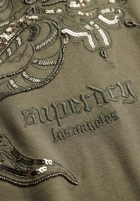 Tela de oliva con texto bordado "superdry los angeles" y diseño floral con cuentas y lentejuelas.