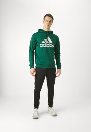 Sudadera verde con el logotipo "adidas" en blanco y tres rayas. Combinada con pantalones jogger negros. La tela parece suave, con un corte atlético y un diseño casual.