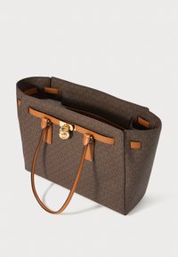 Bolso tote de diseño marrón con patrón de monograma, asas de cuero color crema y detalle de cierre en tono dorado sobre fondo blanco.