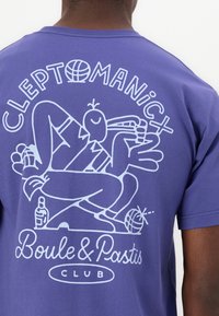 Lilla kortermet t-skjorte med hvit grafikk av en tegneseriefigur, teksten "CLEPTOMANIC Boule & Pastis KLUBB" under.