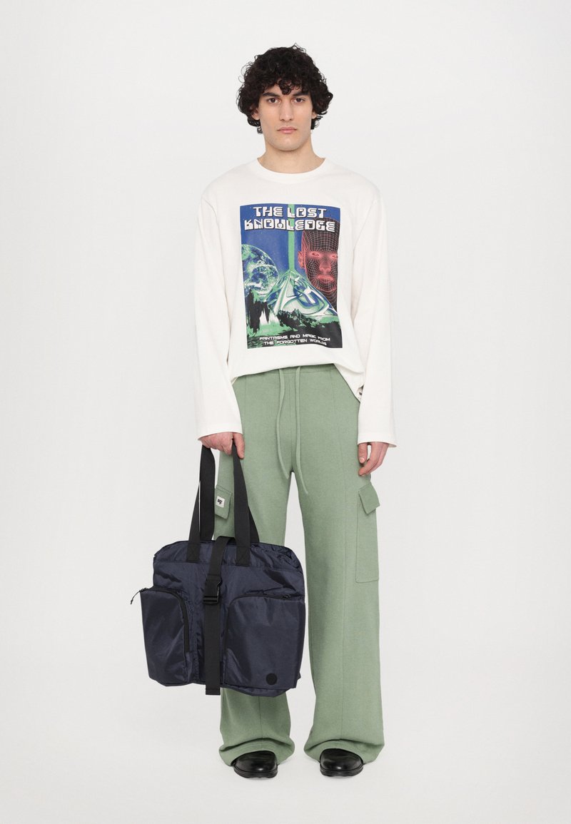 Chemise blanche à manches longues avec un graphisme coloré, pantalon vert à jambes larges et sac à épaule bleu marine avec double poche et bandoulière noire.