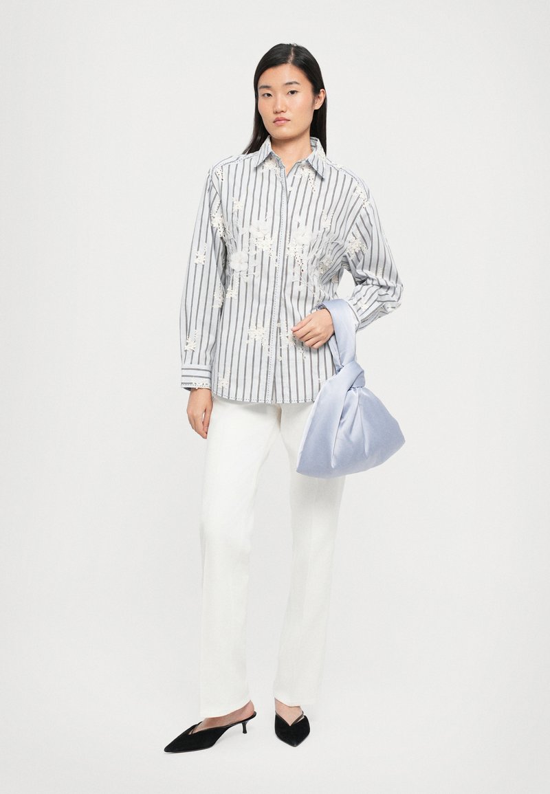 Striped button-up shirt with floral embroidery, paired with white trousers and a soft blue satin bag. Black pointed slip-on shoes complete the look.