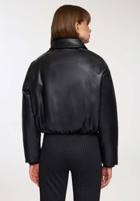 Kobieta o brązowych włosach, ubrana w czarną skórzaną kurtkę i czarne spodnie w prążek, zwrócona plecami w stronę jasnego tła.