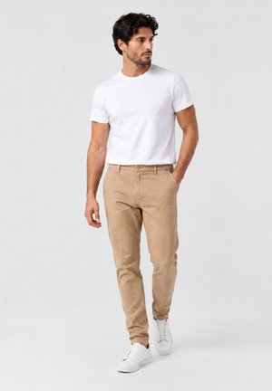 Mann mit dunklem Haar geht, trägt ein weißes T-Shirt, beige Hose und weiße Sneakers vor einem schlichten hellen Hintergrund.
